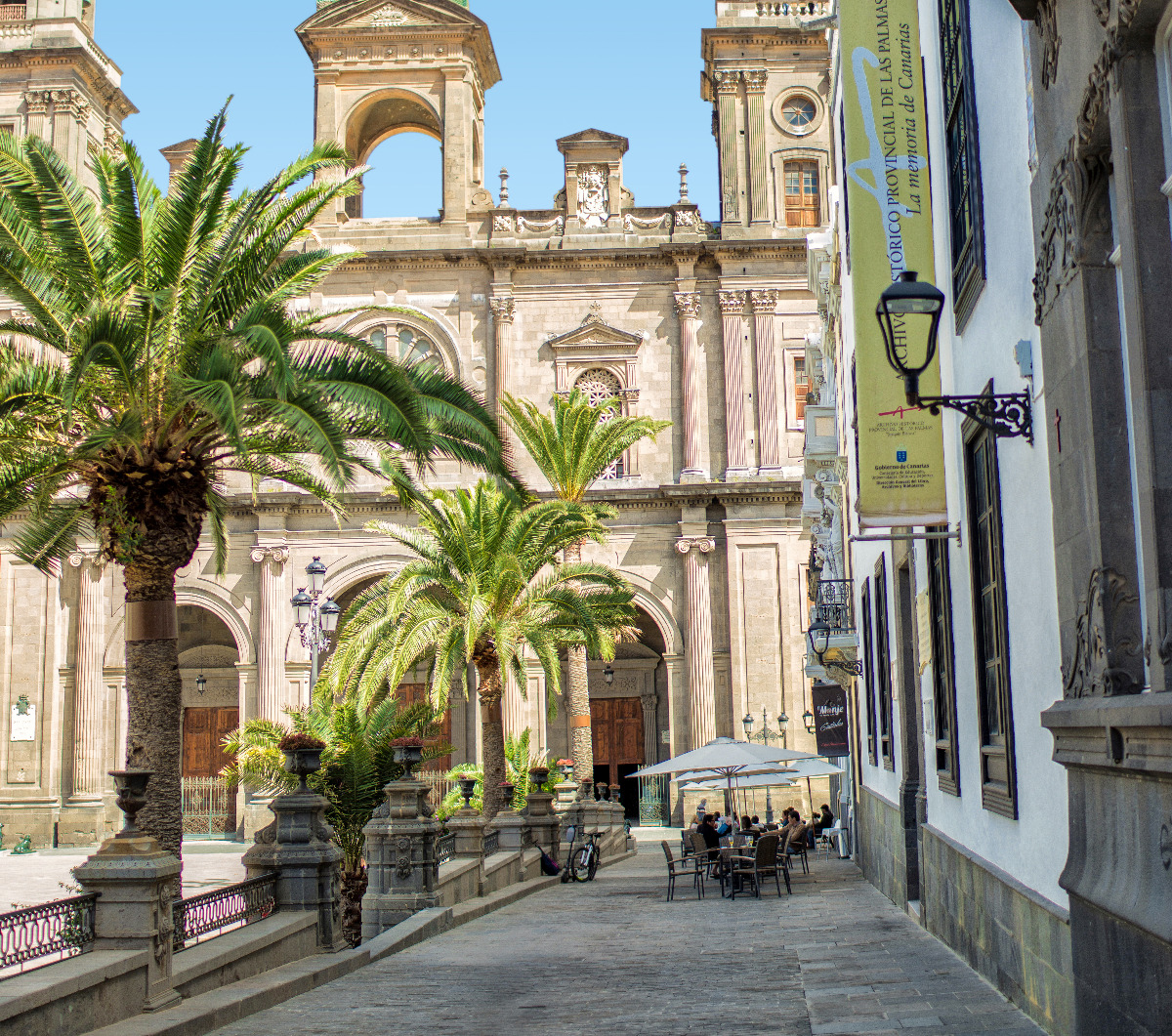 Palmen gesäumte Gasse mit Außencafé, im Hintergrund barocke Kathedrale mit Arkaden und Glockenturm.